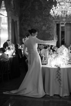 Black and white photo of bride in sleek white gown gesturing at candlelit wedding reception.
