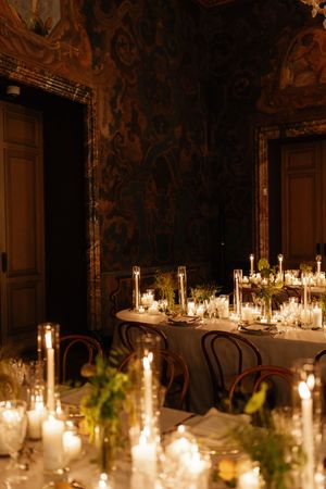 Candlelit wedding reception table in ornate room with dark frescoed walls, bentwood chairs, greenery centerpieces, and glowing tapers.