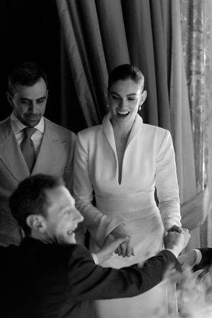 Black and white photo of laughing bride in white blazer greeting guests at reception.