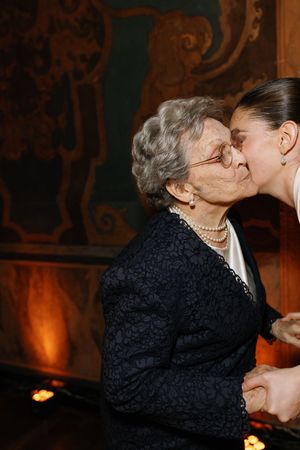 Bride in white pearl-buttoned gown kisses elderly grandmother on cheek at wedding reception.