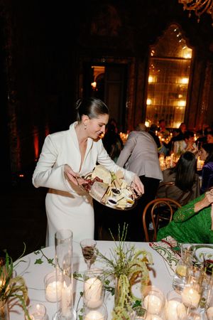 Bride in white suit serves food at candlelit wedding reception dinner table in grand venue.