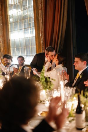 Bride and groom share a kiss at candlelit wedding reception table as guests applaud.