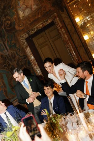 Bride in white suit laughing with wedding guests at ornate candlelit reception dinner.