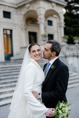 Smiling bride in white coat and veil receives kiss from groom in suit outside grand building.