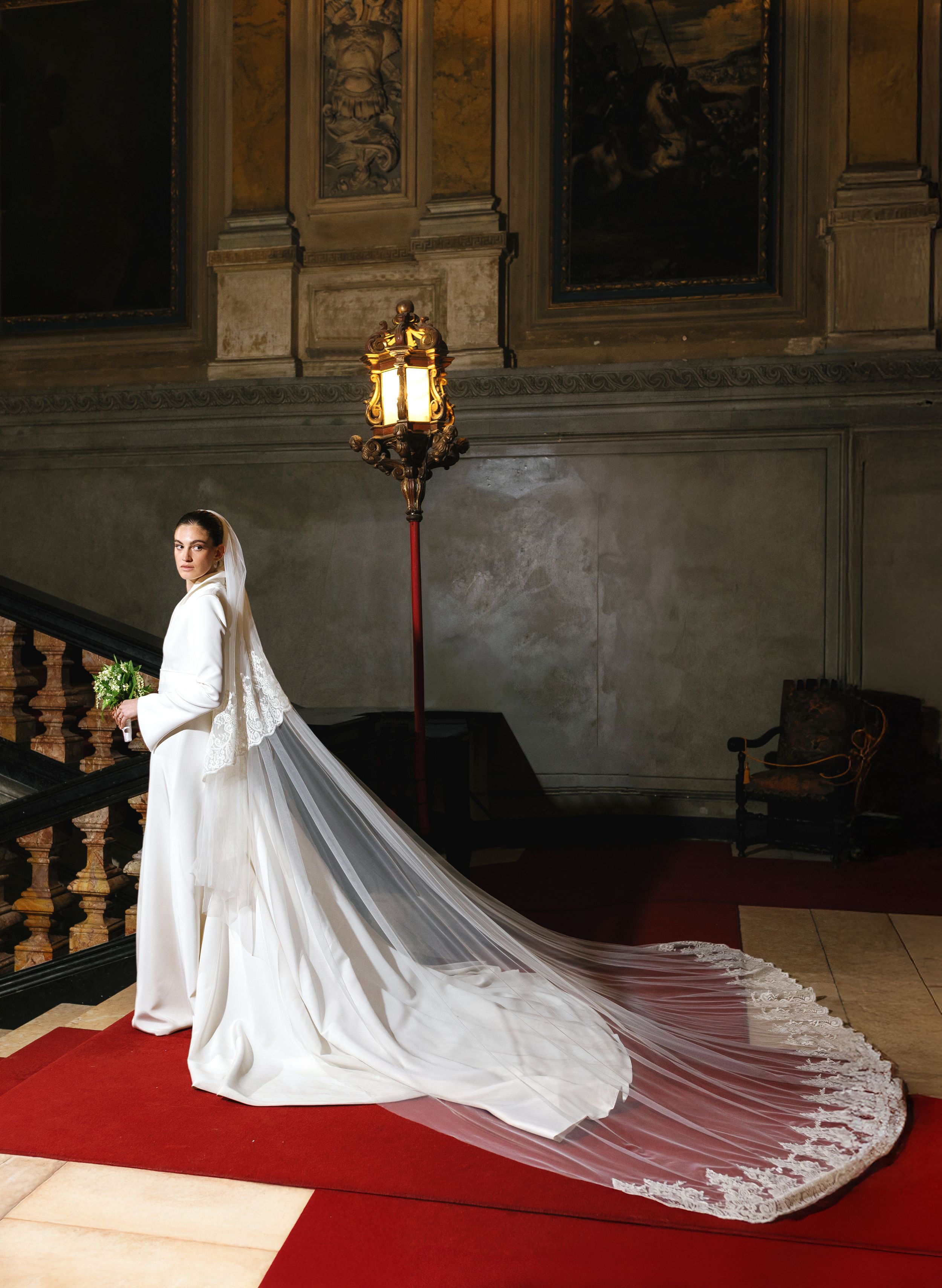 Elegant bride in white gown with long lace cathedral veil on grand staircase.