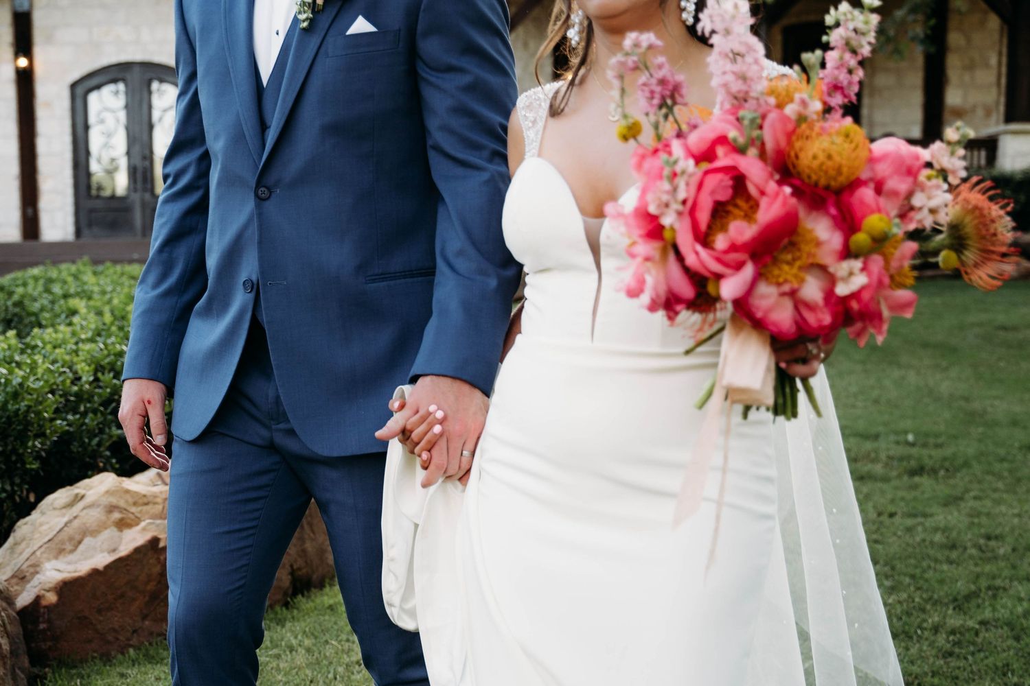 Bride and groom holding hands outdoors, wearing white wedding dress and blue suit, with vibrant pink peony bouquet.