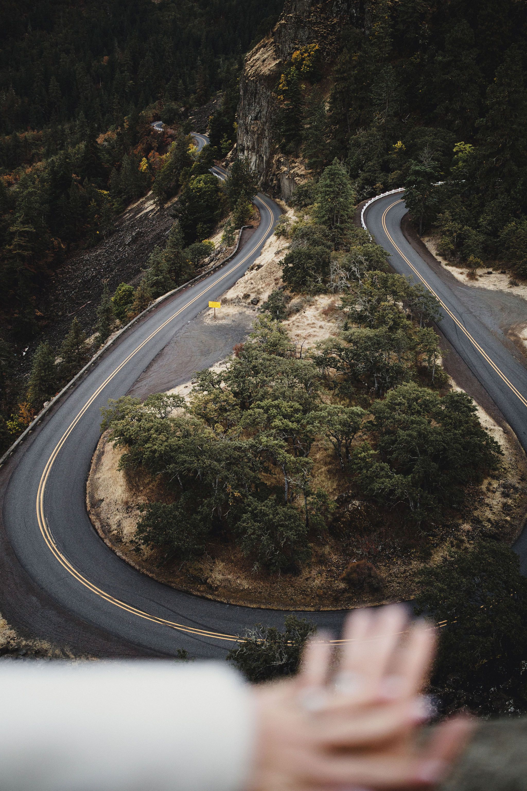 An aerial view of the couples hands with engagement rings rwith the winding mountain road at Rowena Crest in the distance.