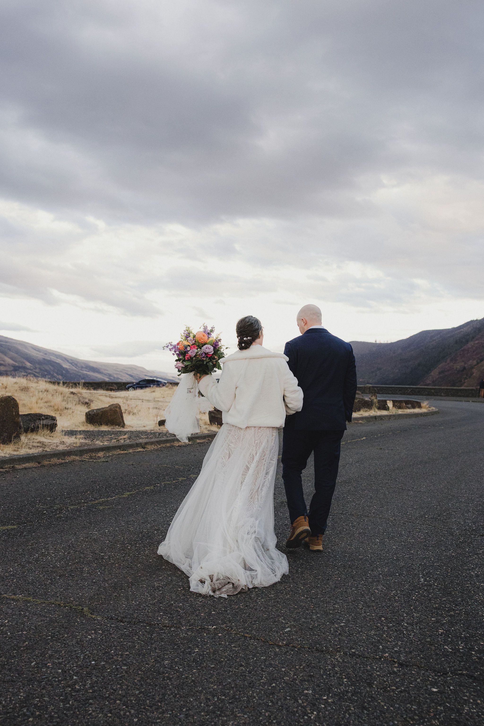 Bride and groom walking hand in hand on a road with mountain views and a colorful bouquet on a cloudy day.