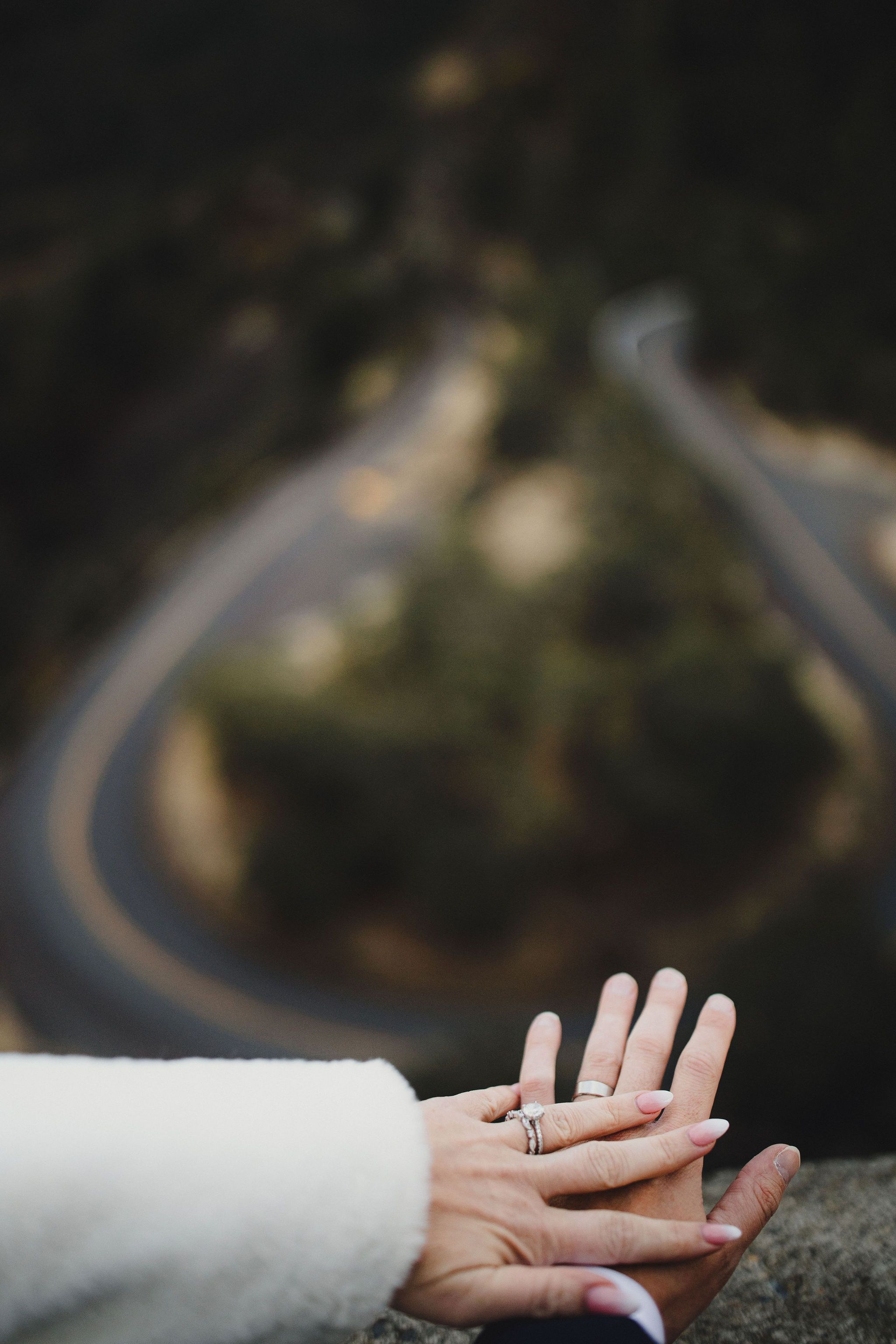 A view of the couples hands with engagement rings rwith the winding mountain road at Rowena Crest in the distance.