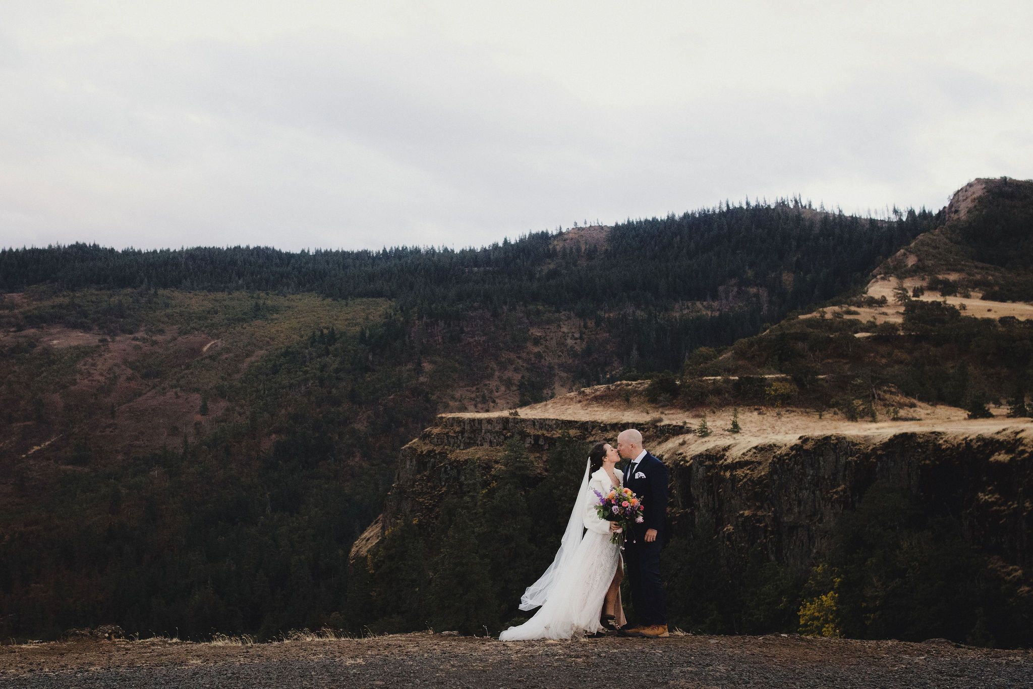 Bride and groom share a romantic embrace on a dramatic clifftop overlooking the Columbia River Gorge in Oregon.