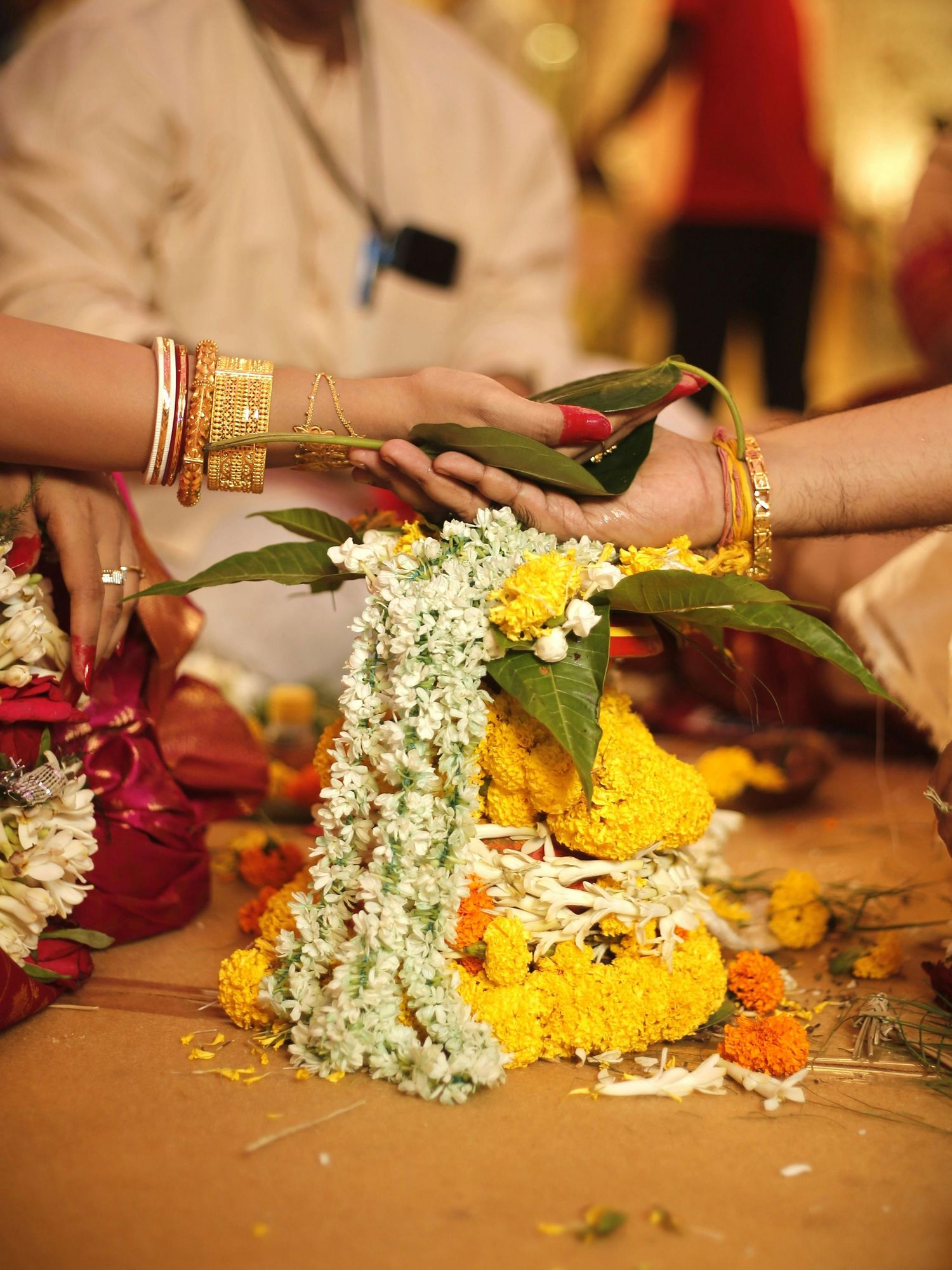 Indian wedding ceremony hands exchanging floral garlands with marigold and jasmine flowers, bride wearing gold bangles.