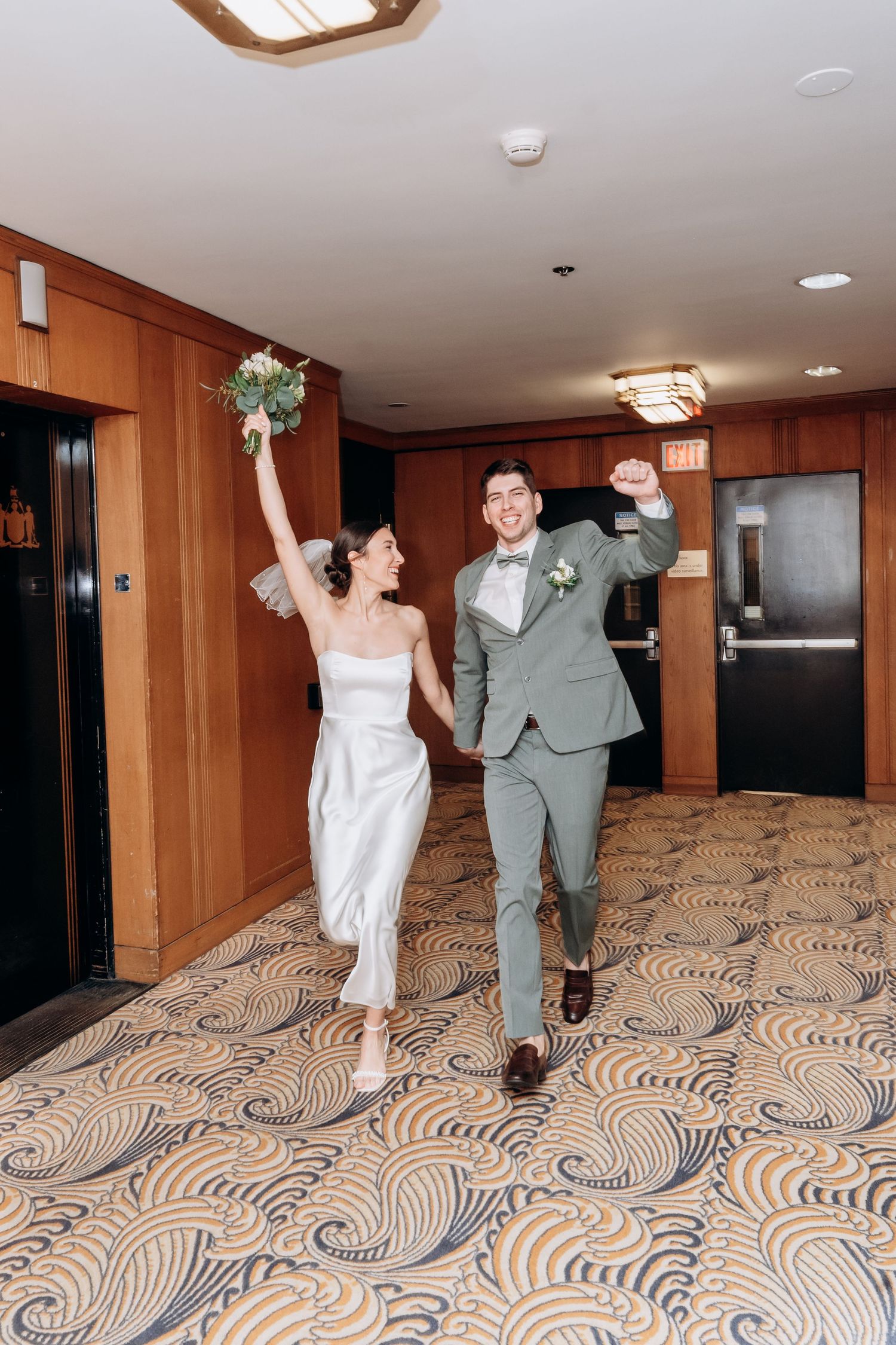 A joyful couple celebrates in a hotel hallway with wood paneling and patterned carpet after their wedding ceremony.