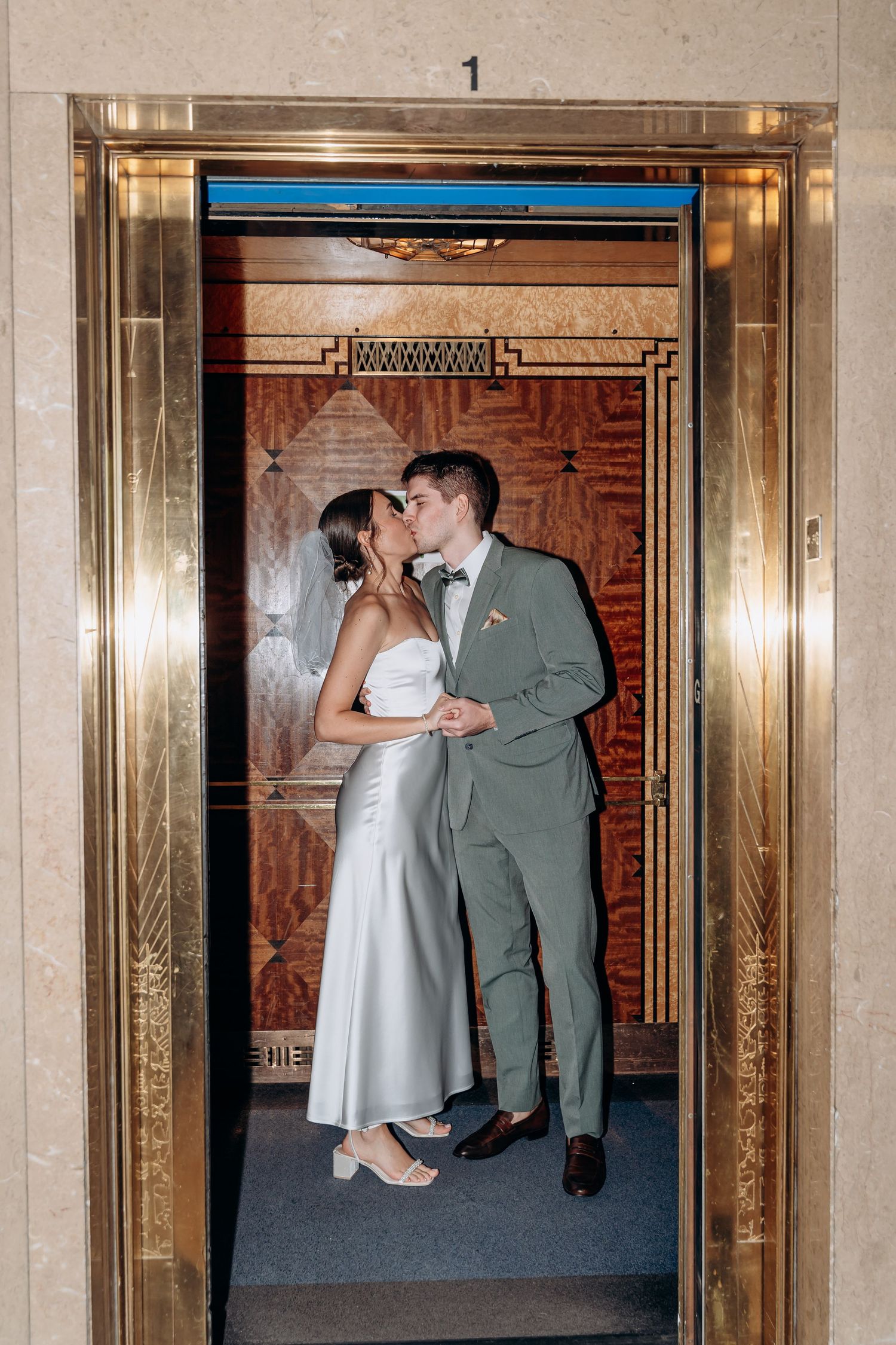A couple shares a romantic kiss in front of a vintage elevator with ornate wood paneling and mirrored walls.