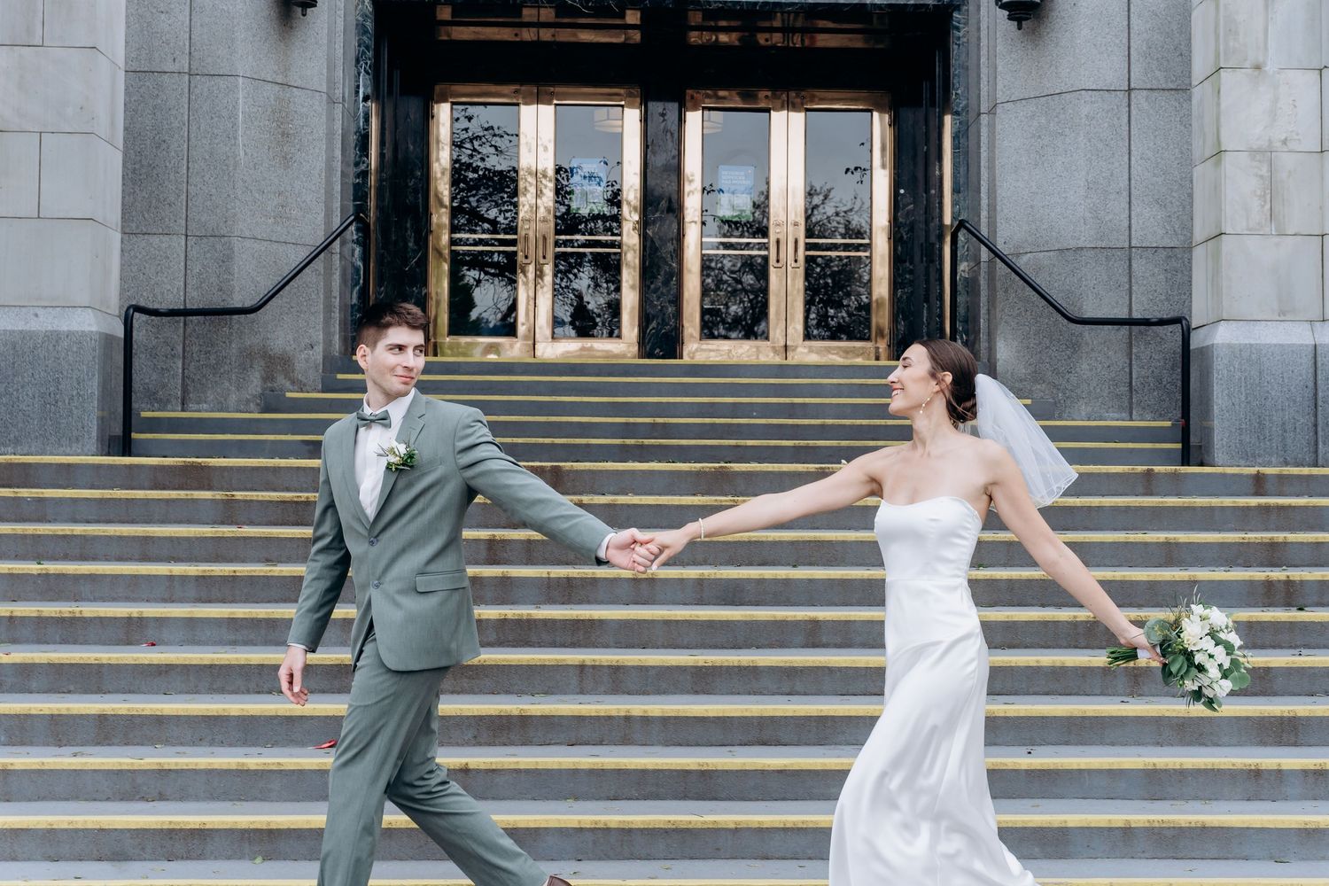 A bride and groom hold hands while walking apart on stone steps outside a grand building with columns.
