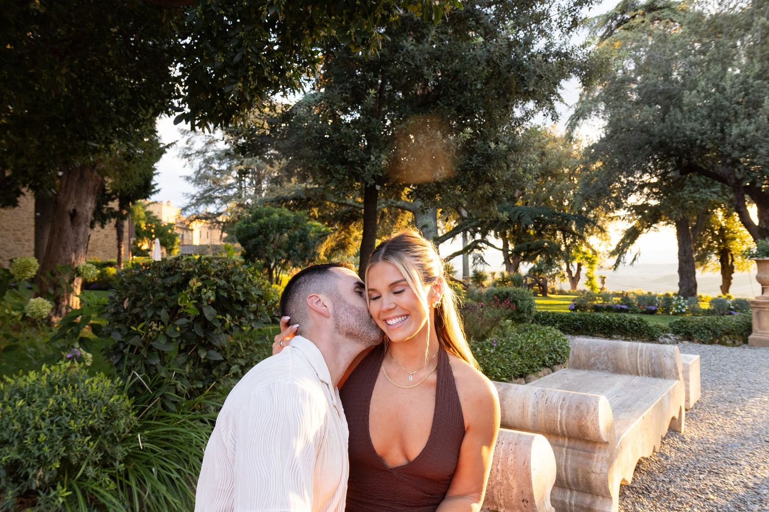 A romantic couple shares a tender moment at sunset in a lush garden setting with stone benches.
