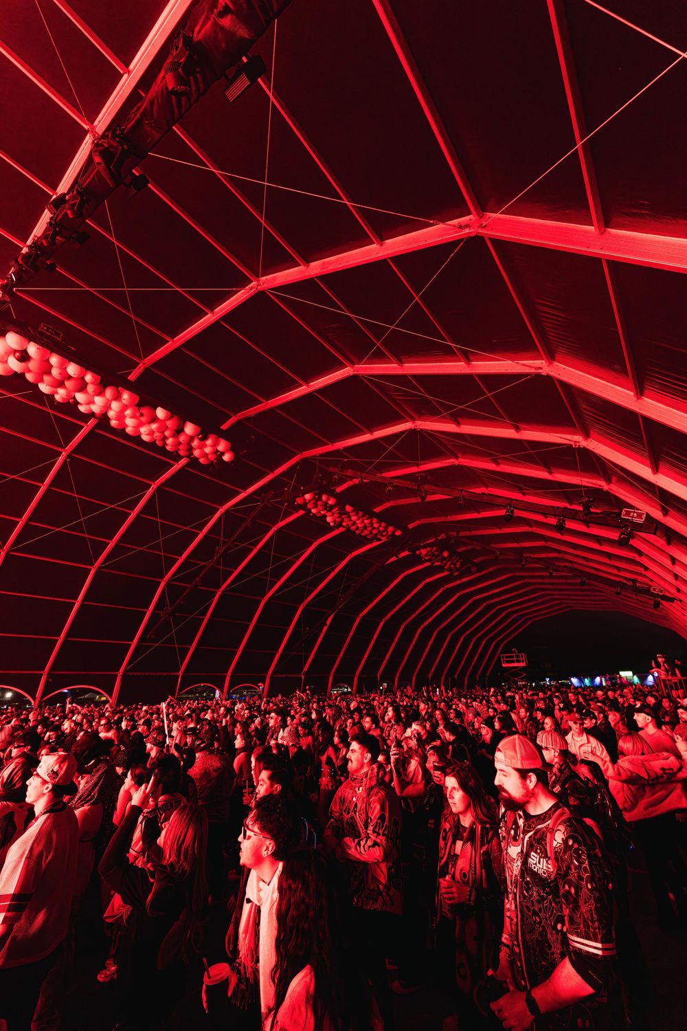 A crowd of concert-goers bathed in red lighting under an arched tunnel structure at a music event.