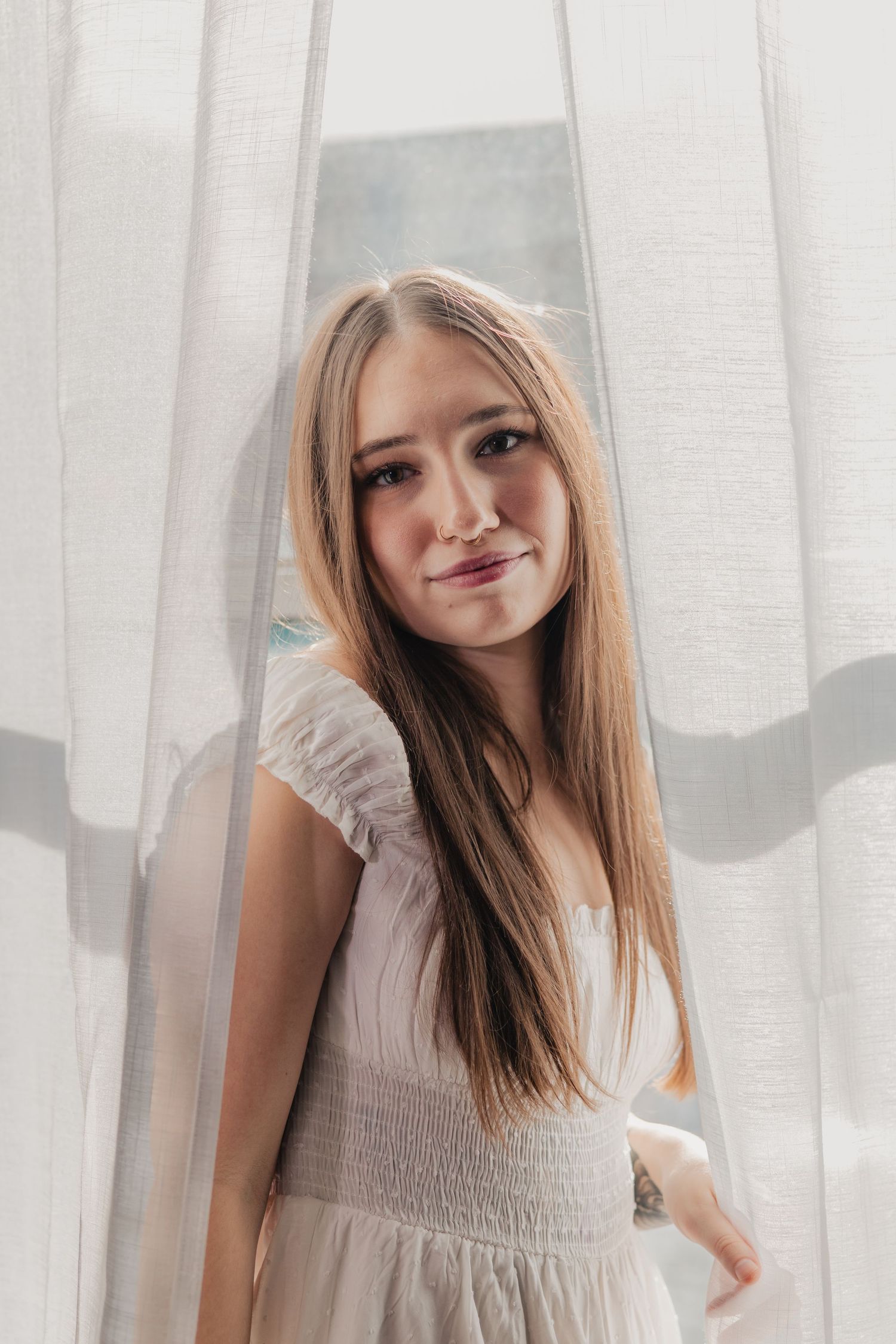 A person with long straight brown hair poses in front of white curtains wearing a light colored sleeveless dress.