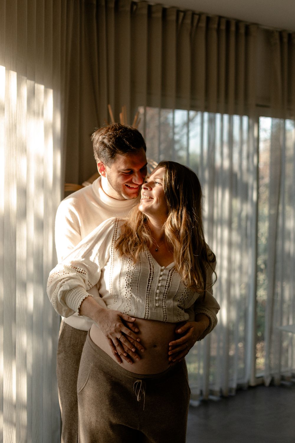 A couple shares an intimate moment in a sunlit hallway with white curtains creating a dreamy atmosphere.