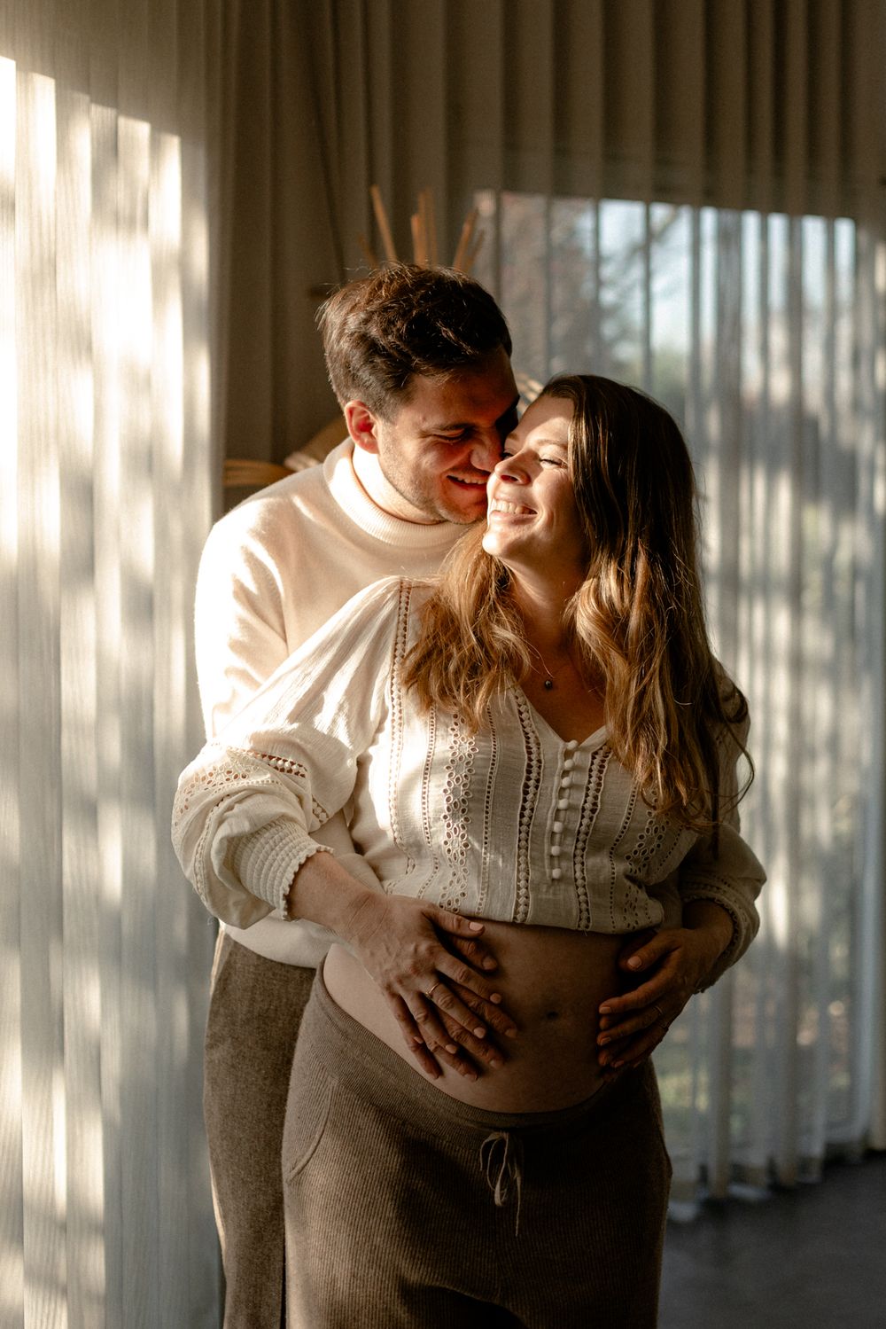 Romantic couple in white sweaters embracing in sunlit room with window shadows creating dreamy atmosphere.