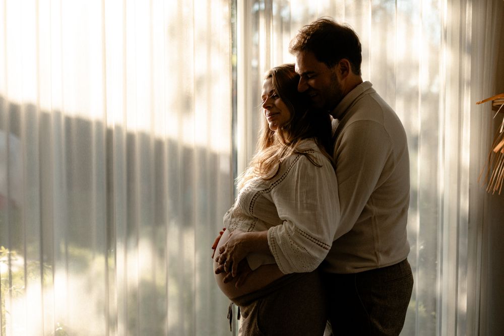 A silhouetted couple embraces near a sunlit window with sheer curtains creating a dreamy, romantic atmosphere.