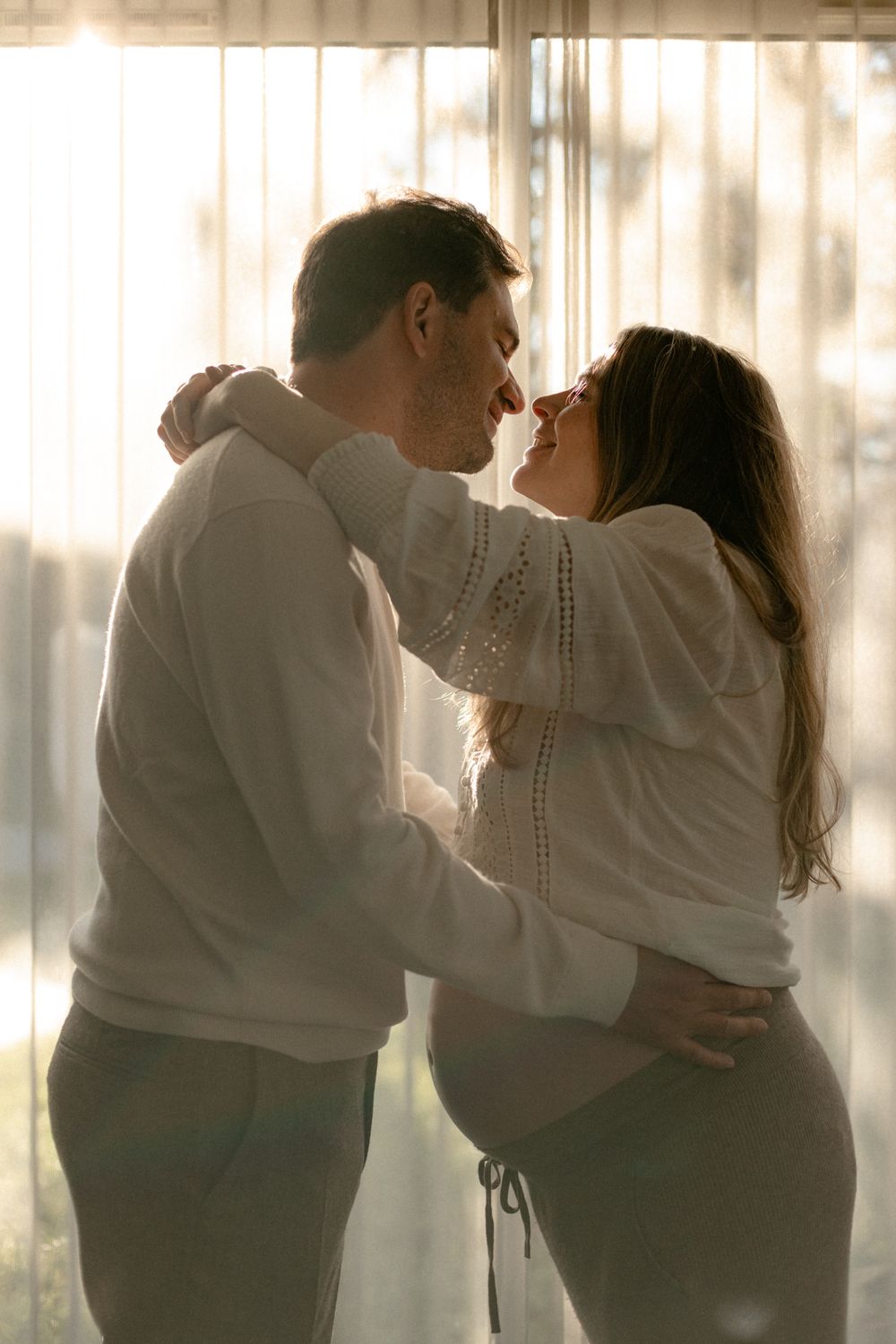 Couple sharing intimate moment in soft window light wearing matching white knit sweaters.