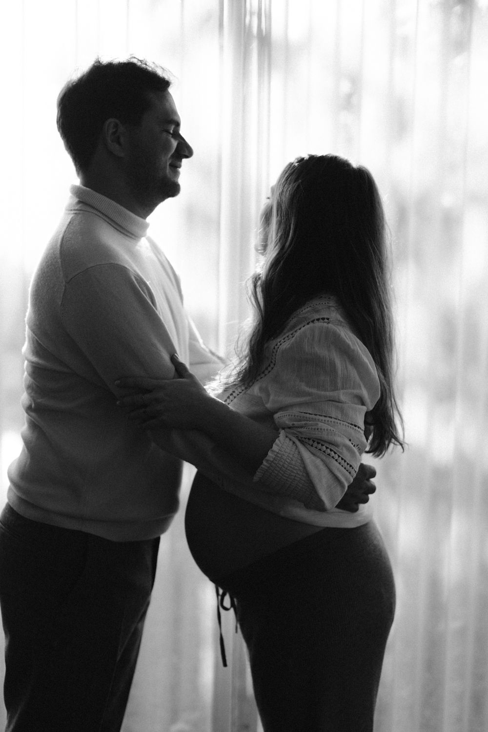 Black and white silhouette of couple embracing by window, showing pregnant woman's belly in profile.