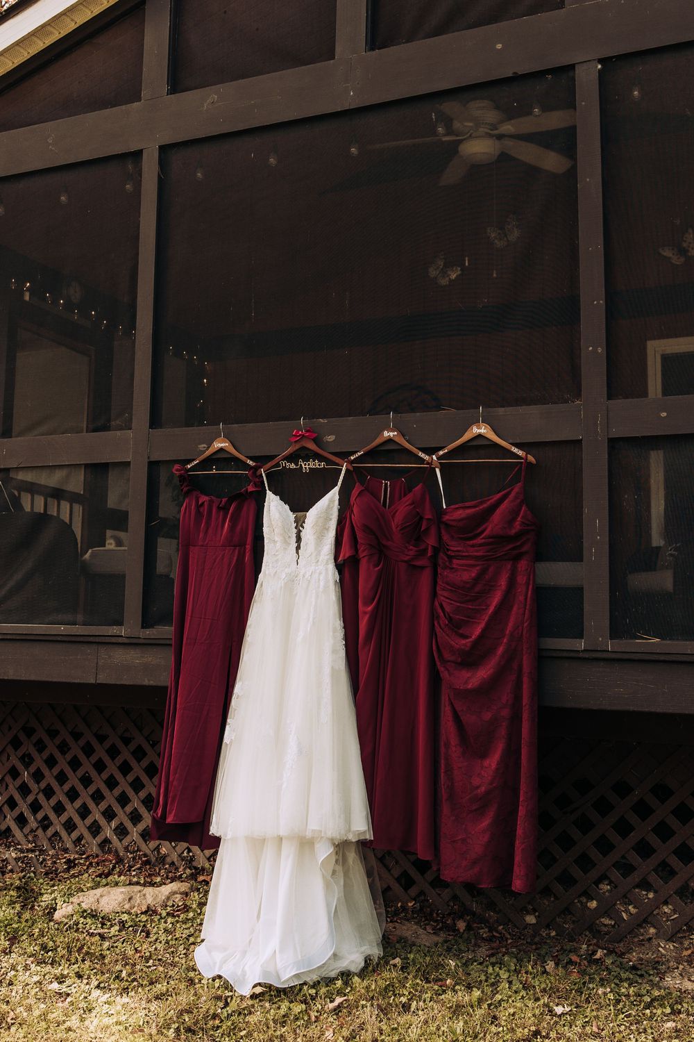 White wedding gown and three burgundy bridesmaid dresses hanging outside a rustic screened porch.