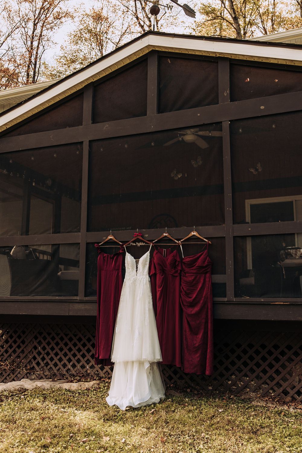 Bridal gown flanked by deep red bridesmaid dresses displayed on hangers outside a wooden screened cabin.