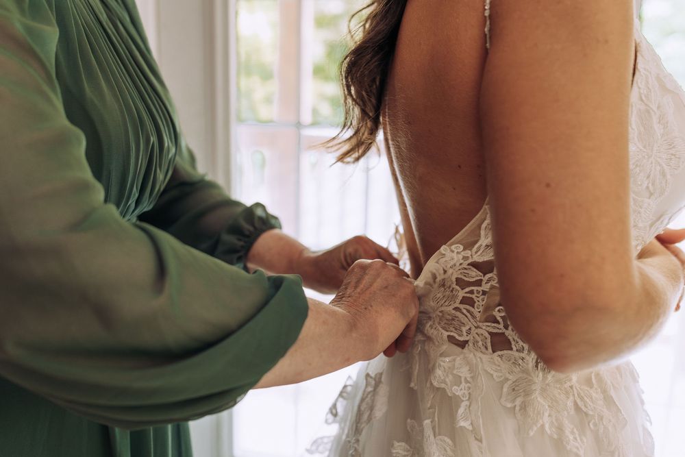 Close-up of hands fastening the back buttons of a lace bridal gown near a bright window.