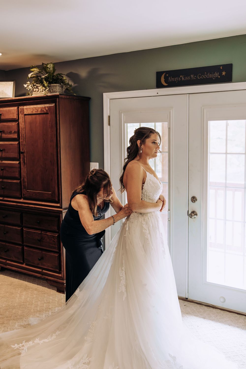 Mother helps bride button her lace wedding dress near French doors in a green-walled room.