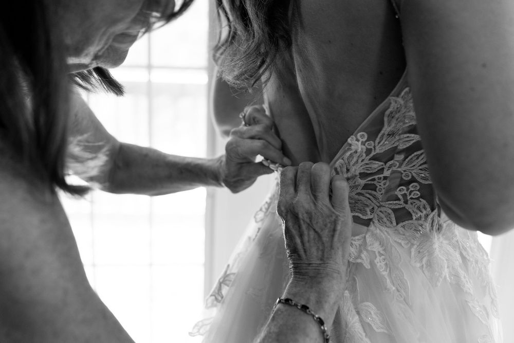 Black and white photo of hands carefully buttoning the intricate lace back of a wedding dress.