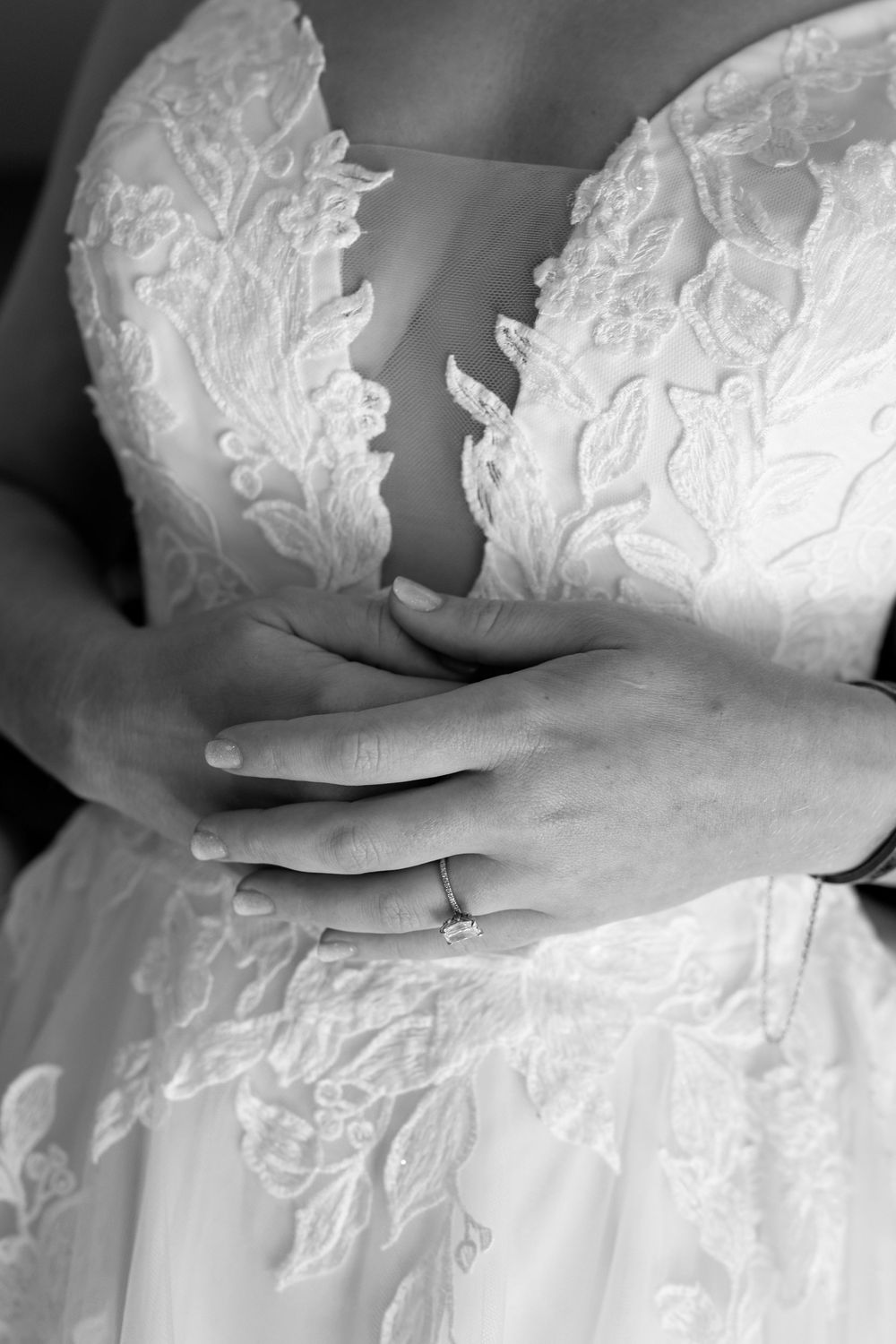 Black and white close-up of bride in lace gown with diamond engagement ring resting on her hands.