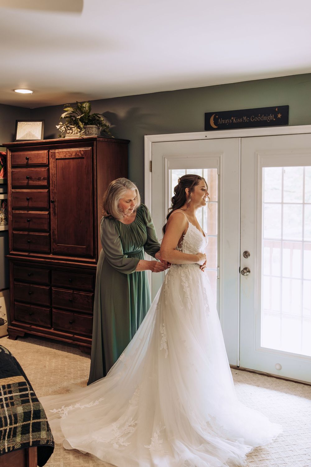 Older woman in teal dress helping bride with her lace wedding gown train near French doors.