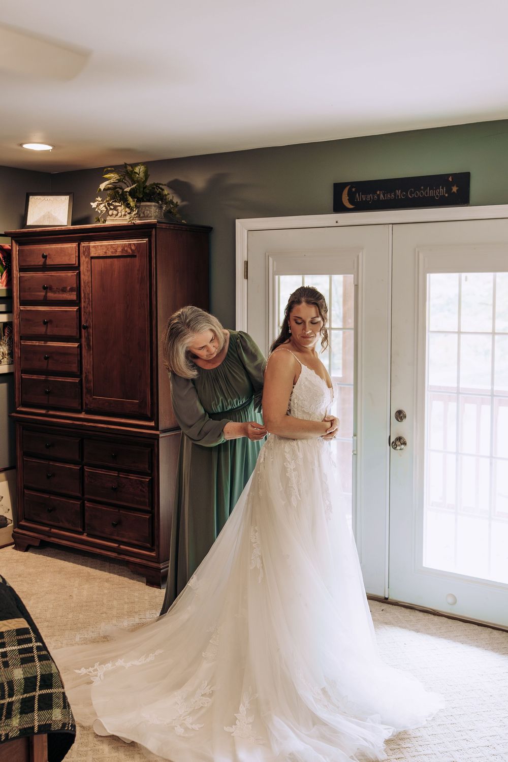 Mother in green dress assists smiling bride adjusting her lace wedding dress in a cozy bedroom.