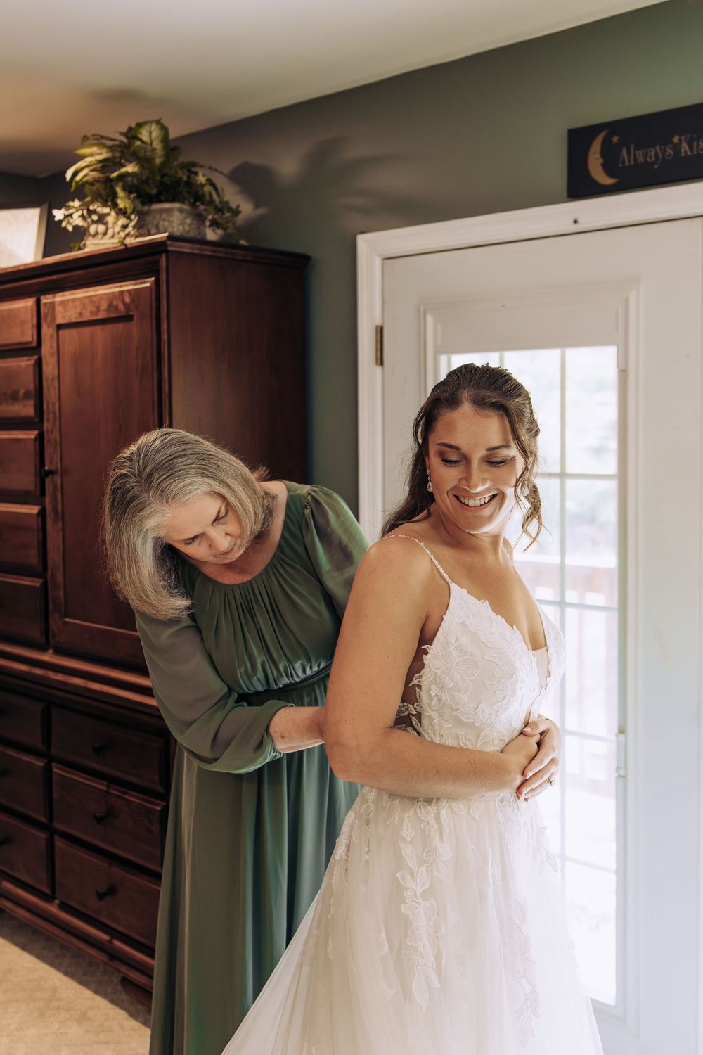 Bride smiles joyfully as her mother helps fasten her lace wedding gown beside a wooden armoire.