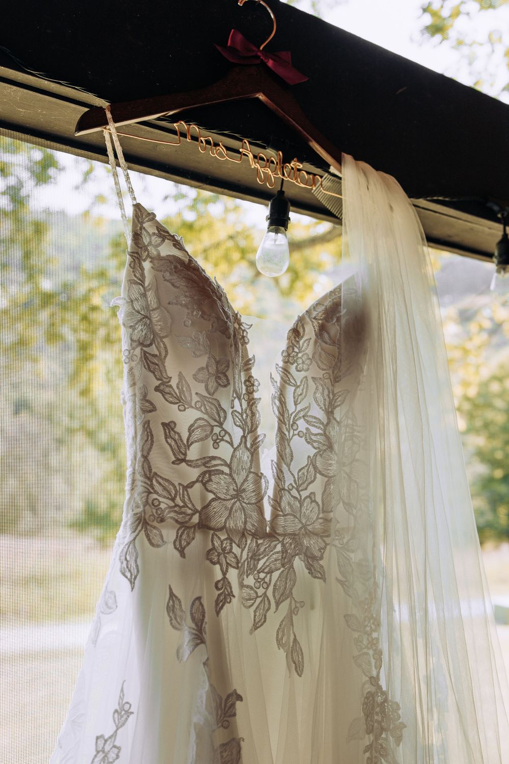 Close-up of an intricate lace wedding dress hanging on a personalized hanger in a window with veil.