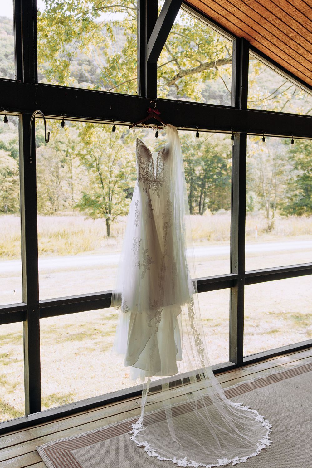 Elegant lace wedding dress with long cathedral veil hanging in a large window overlooking a meadow.