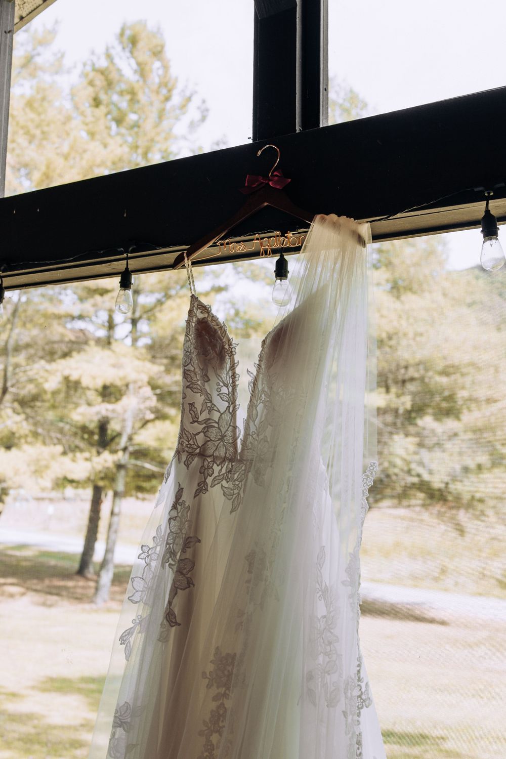 Beautiful lace wedding gown with flowing veil hanging on a personalized hanger in a sunlit window.