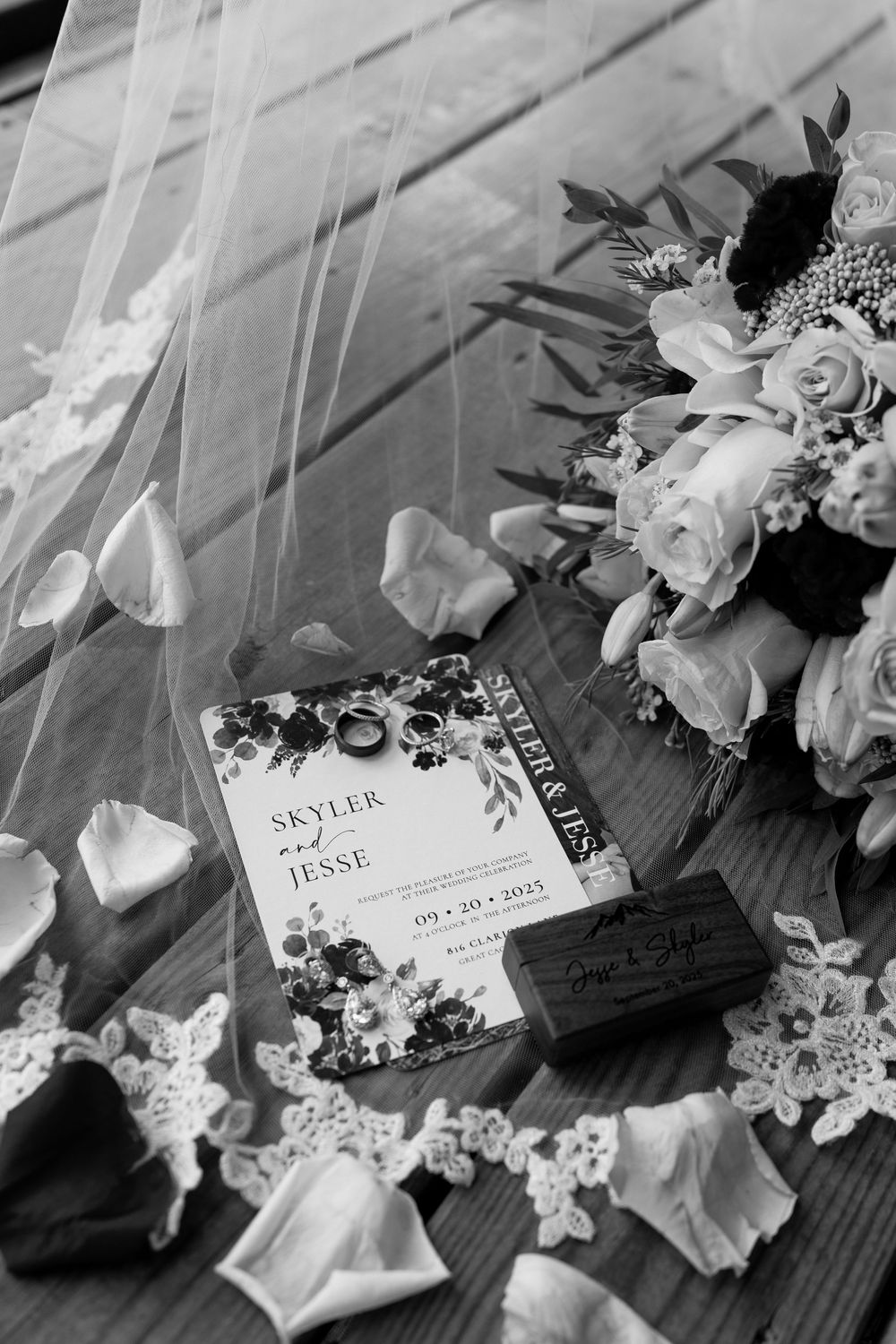 Black and white flat lay of wedding invitation for Skyler and Jesse with rings, bouquet, and lace veil on wood.