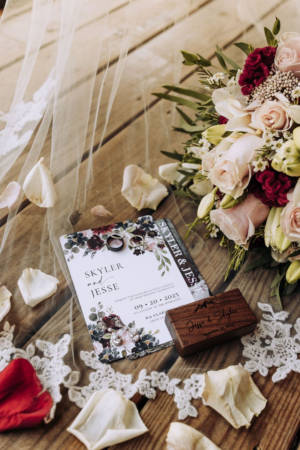 Wedding flat lay featuring floral invitation for Skyler and Jesse, bouquet, ring box, and rose petals on wood.