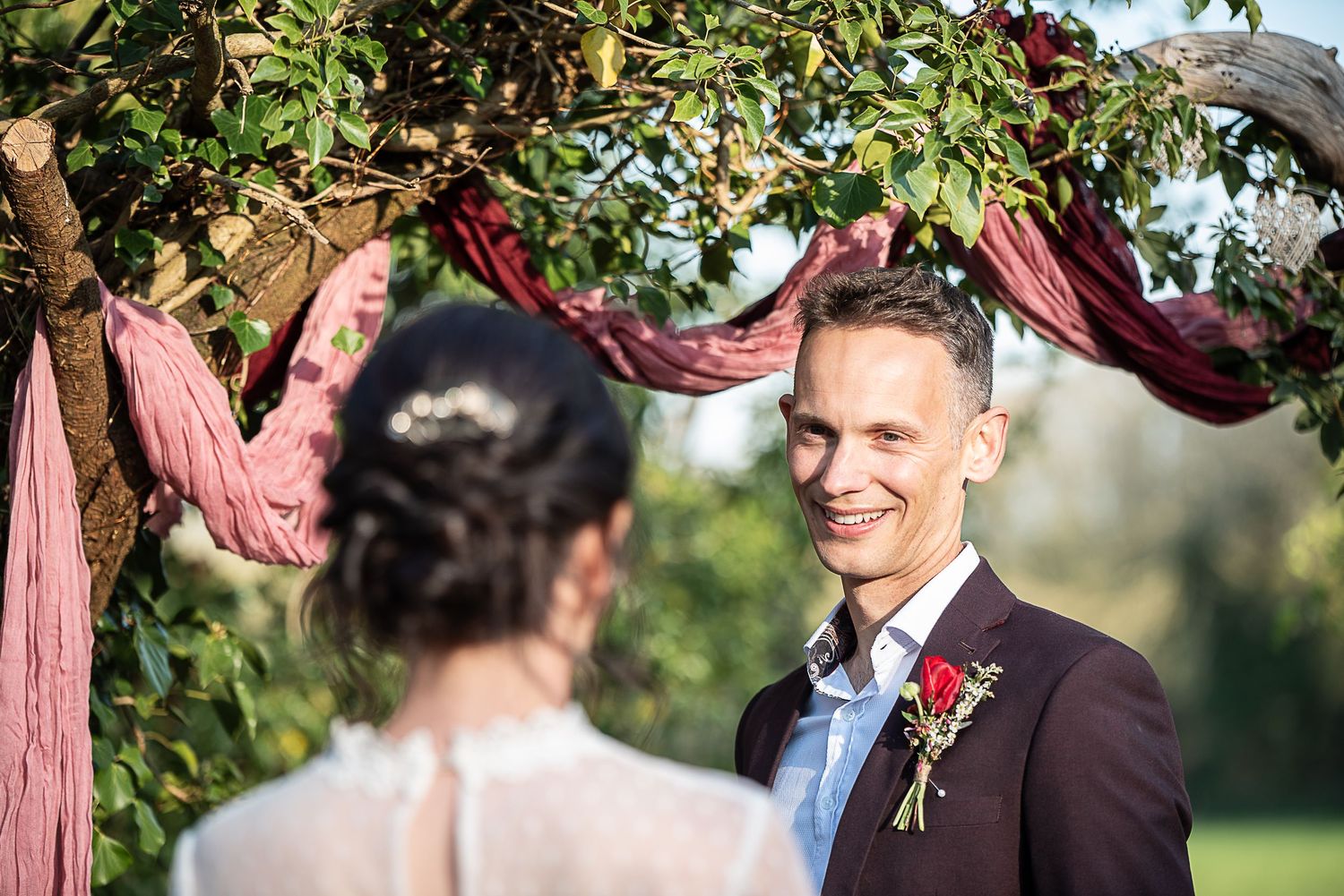 Groom in sunshine smiles as bride walks towards him. He wears a red button hole and is surrounded by greenery at Nature's Edge Cabin