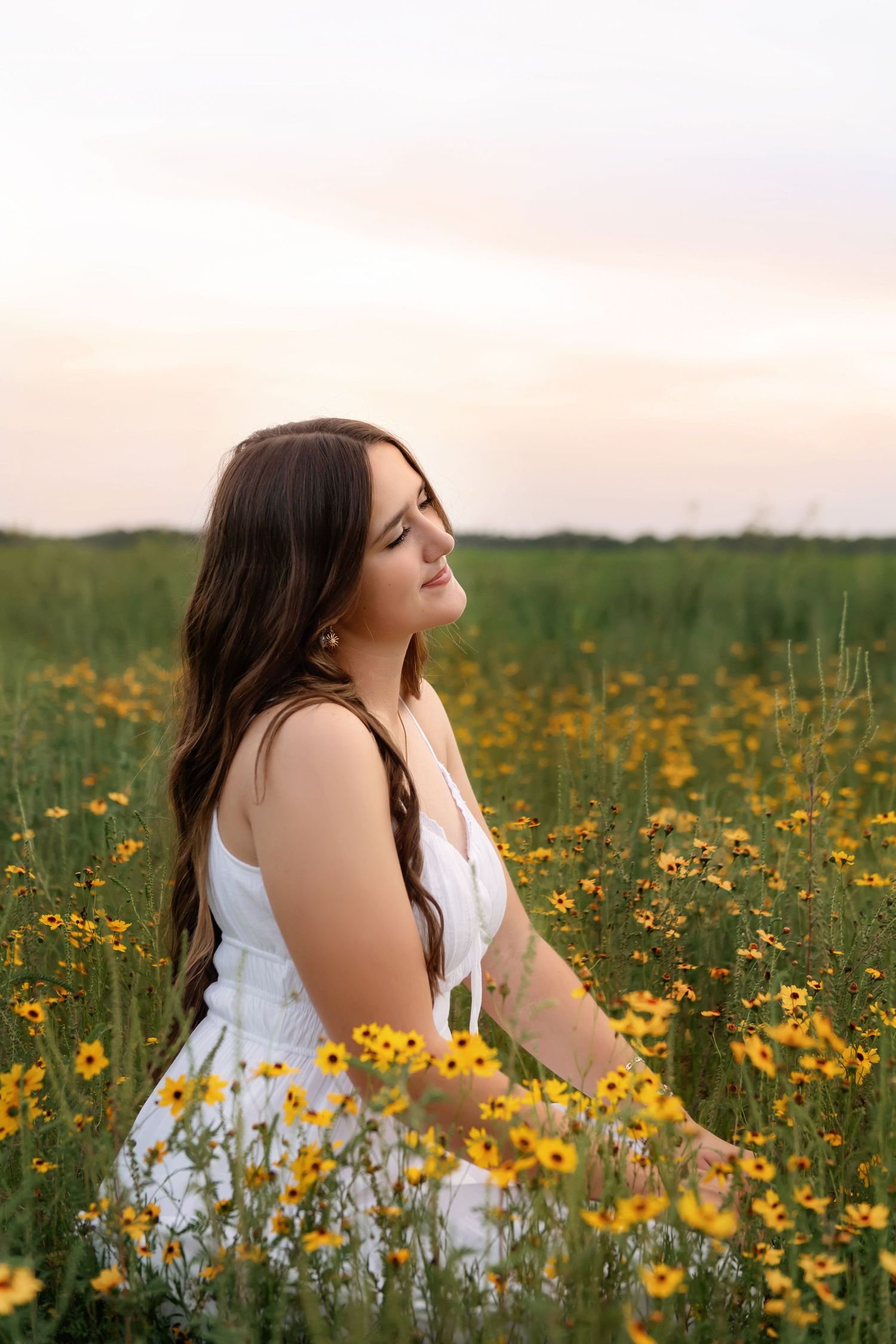 Young woman in white dress sitting peacefully among vibrant yellow wildflowers at sunset.