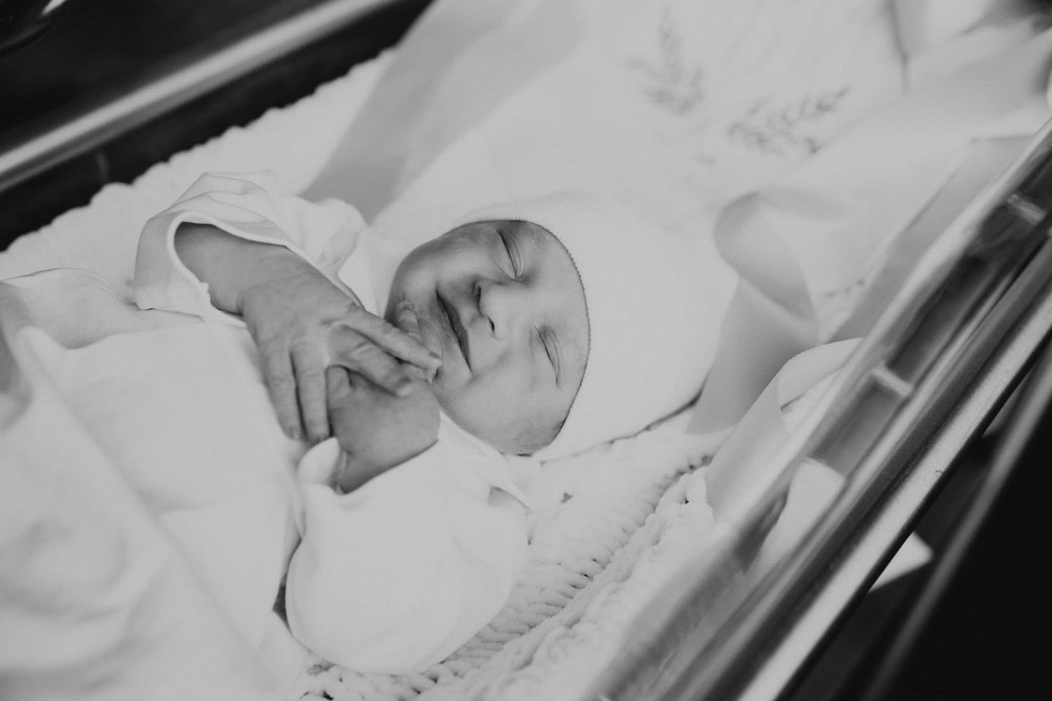 Black and white close-up of a peaceful newborn baby wrapped in white blanket in a hospital bassinet.