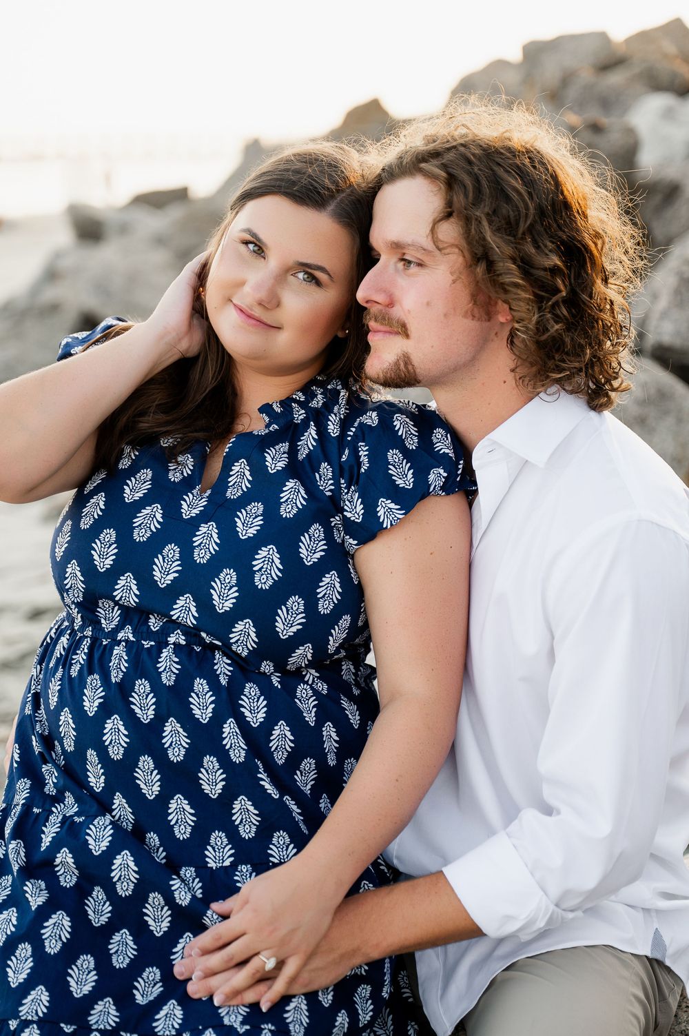 Couple in navy patterned dress and white shirt embracing on rocky beach.