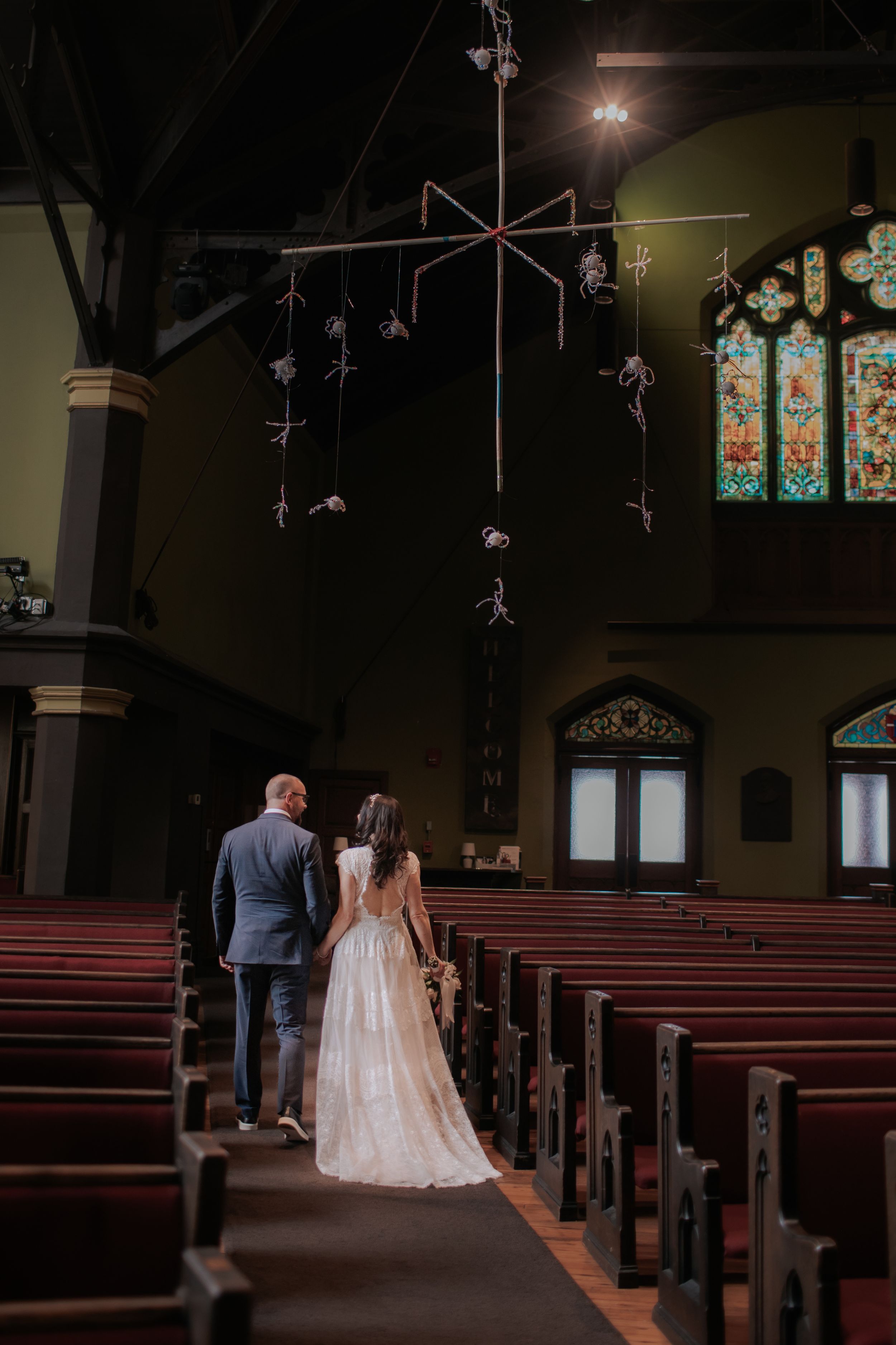 A couple walks down the aisle of a church with beautiful stained glass windows and hanging decorative elements.