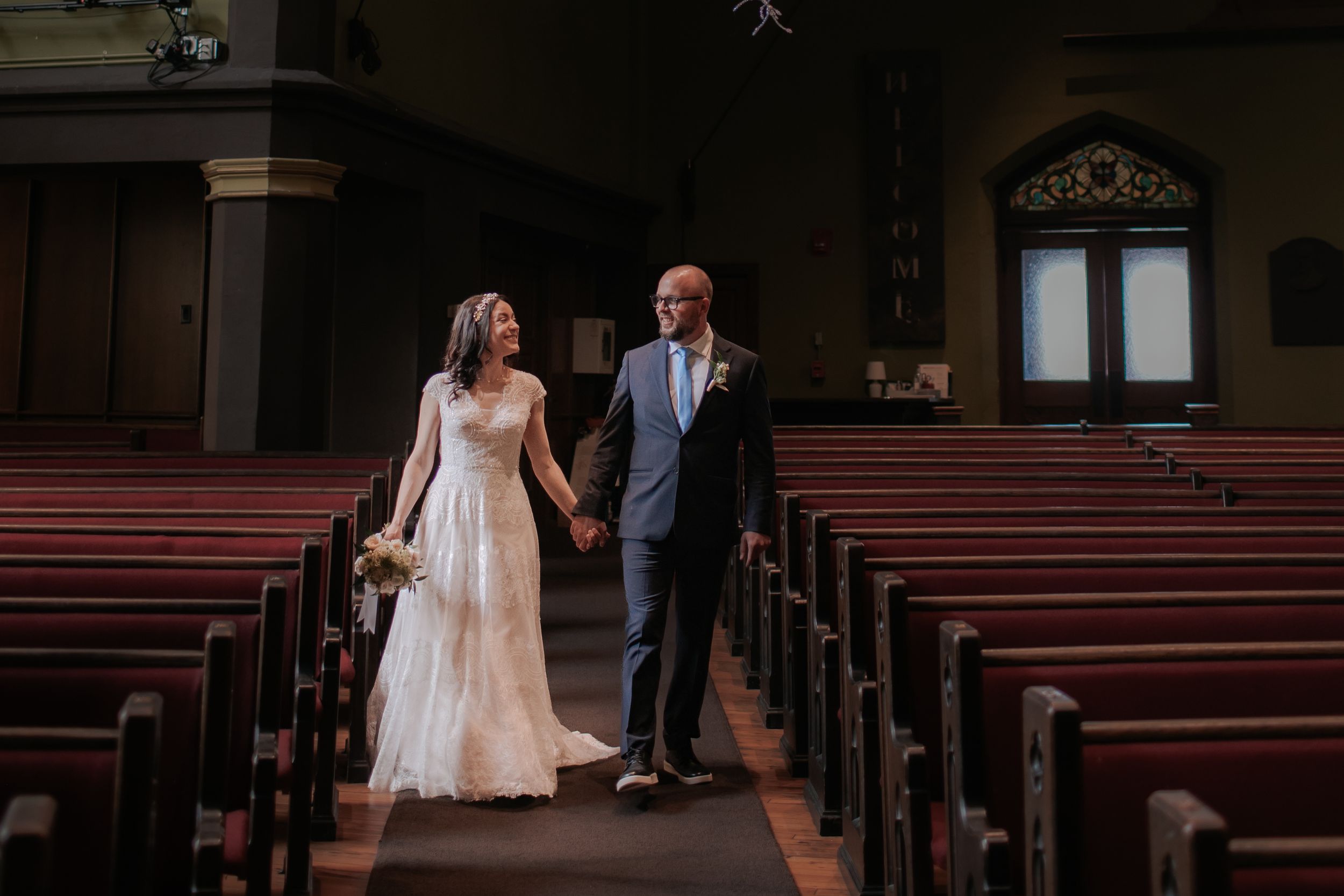 A couple walks down the aisle of a historic church with wooden pews and ornate architectural details.