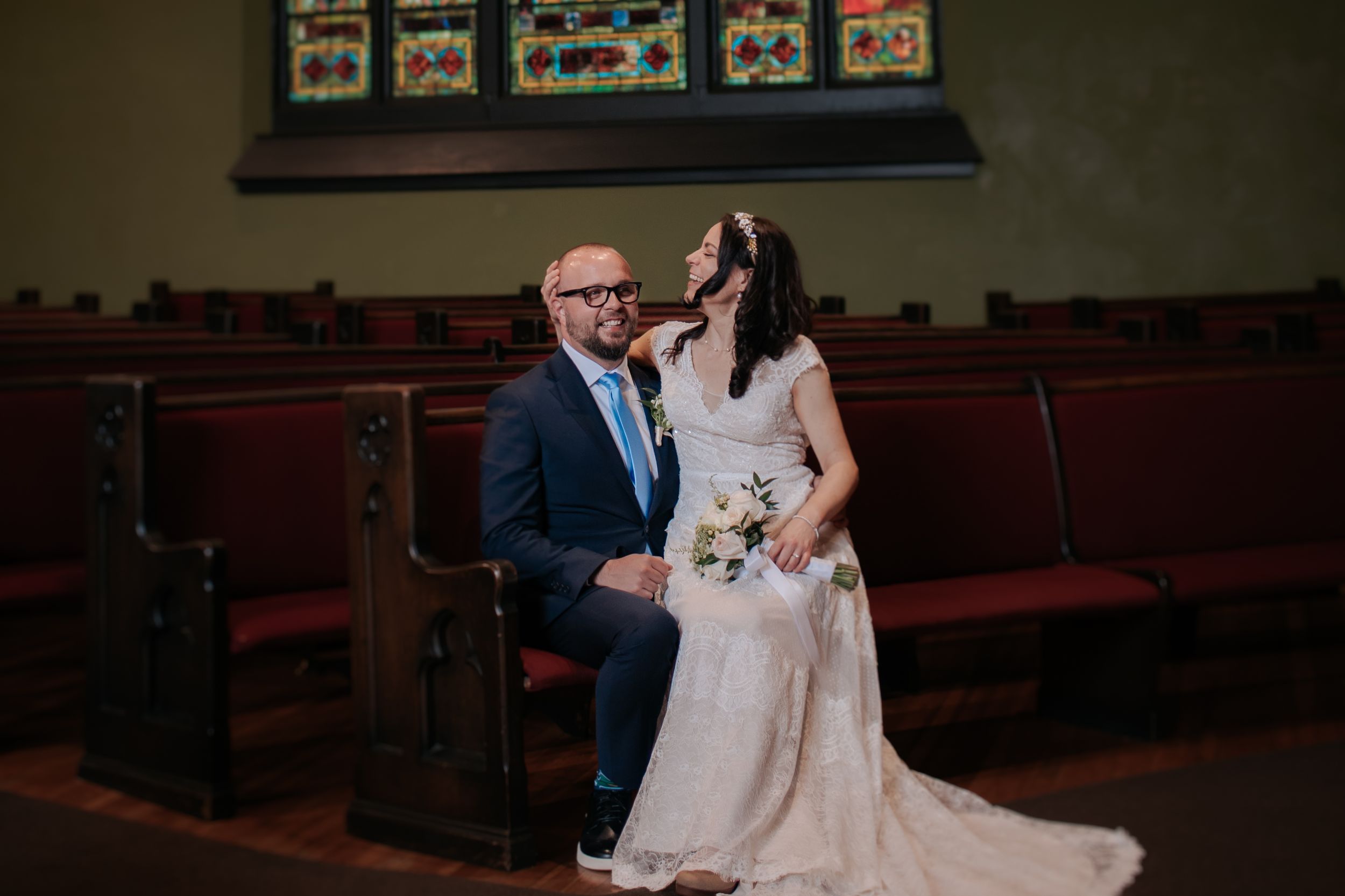 A couple sits together in an old church pew, the bride wearing a flowing white gown and holding a floral bouquet.