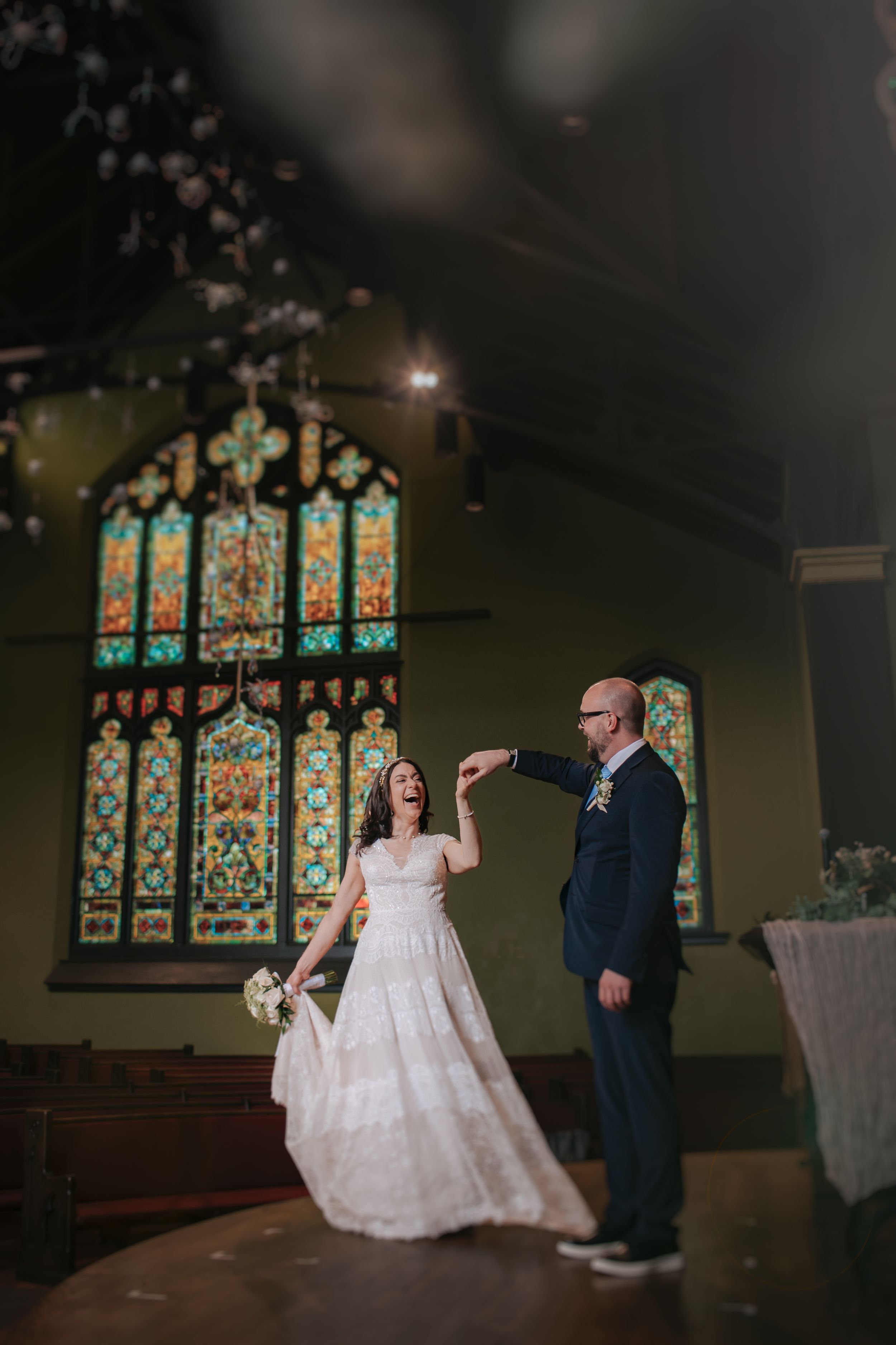 A couple shares a romantic dance moment in a church with beautiful stained glass windows in the background.