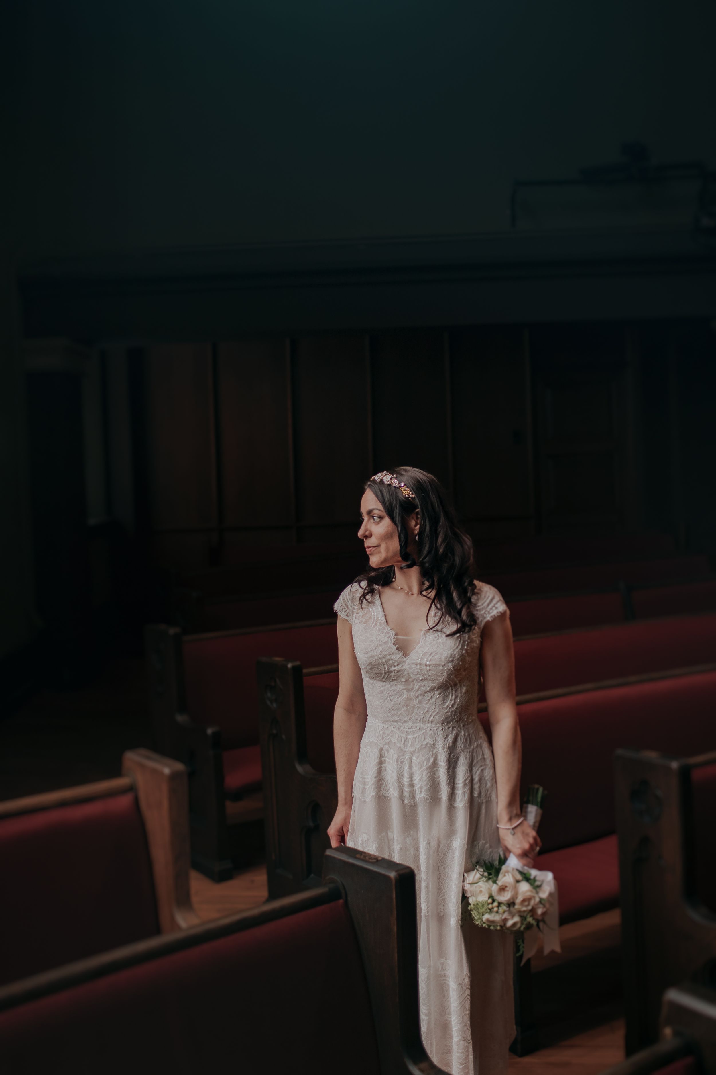 A bride in a white lace dress stands alone in a dimly lit church pew holding a floral bouquet.