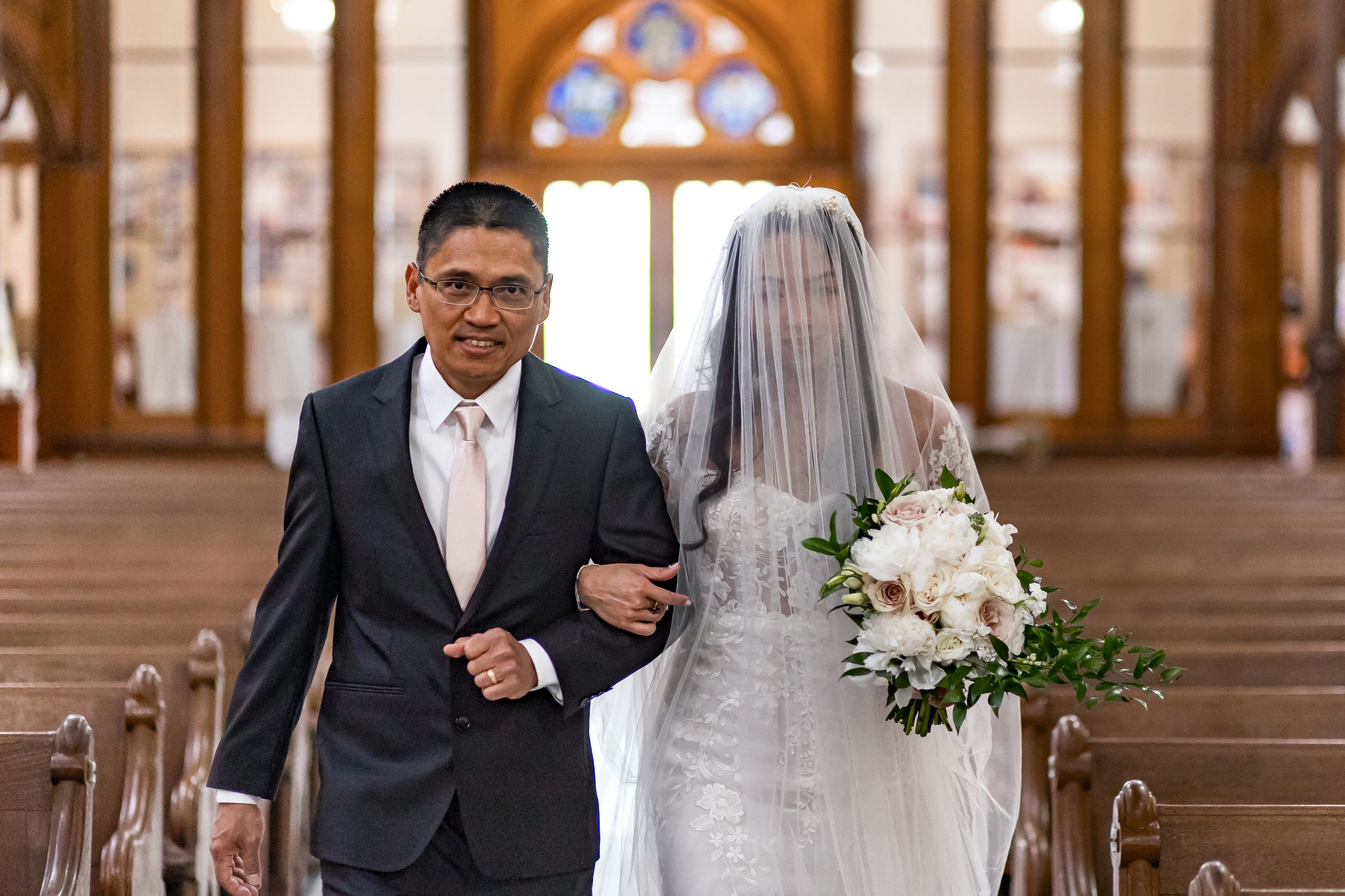 A wedding couple walks down the aisle of an elegant church with arched windows and ornate architectural details.